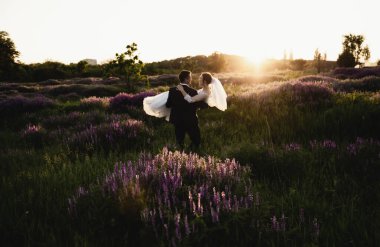 Yeni evliler gün batımında çiçek açan bir tarlanın ortasında duruyorlar, damat gelini kollarında tutuyor, düğün gününde mutlu çift, gelin ve damadın arka planında çiçek açan tarla, gelin ve damat gün batımında sarılıyor. Mutlu gelin ve damat gülümsemesi.
