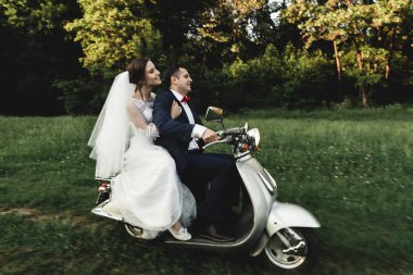 Düğün damadı ve gelin motosiklet sürüyor, eğleniyor, gülümsüyor düğün çifti gün batımında şehir dışındaki yolda scooter 'a biniyor, düğün çifti scooter' a biniyor, yeni evliler dekore edilmiş scooterla eğleniyor, bulanık yeni evliler.