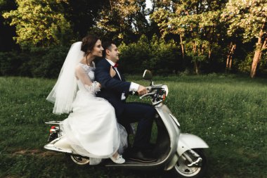 Düğün damadı ve gelin motosiklet sürüyor, eğleniyor, gülümsüyor düğün çifti gün batımında şehir dışındaki yolda scooter 'a biniyor, düğün çifti scooter' a biniyor, yeni evliler dekore edilmiş scooterla eğleniyor, bulanık yeni evliler.