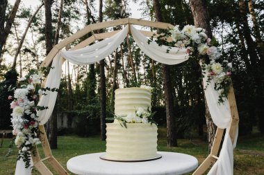 Beyaz ranzalı düğün pastası bir masanın üzerinde duruyor, gelin bir düğün pastası kesiyor, çiçekçilerle süslenmiş düğün pastası, düğün gününün sonunda bir düğün pastası kesiliyor, dışarıda pasta var.