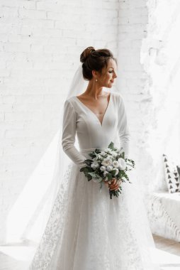 Güzel bir çiçek buketiyle beyaz bir stüdyo duvarına poz veren genç bir gelin, güzel bir gelinin muhteşem gelinliği, şık bir gelinlik, bir buket çiçek.