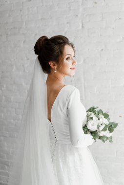 Güzel bir çiçek buketiyle beyaz bir stüdyo duvarına poz veren genç bir gelin, güzel bir gelinin muhteşem gelinliği, şık bir gelinlik, bir buket çiçek.