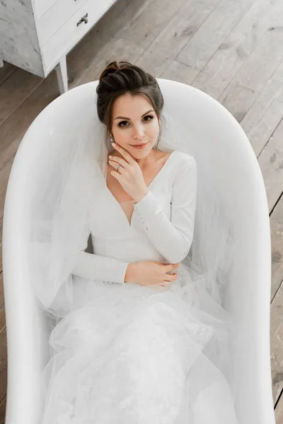 Genç gelin banyoda yatıyor, düğün günü gelini, gelinlikli güzel kız beyaz bir banyoda yatıyor, gelin portresi, düğün fotoğrafı çekimi, gelinlik, gelin elleriyle yüzüne dokunuyor.