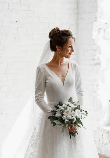 Güzel bir çiçek buketiyle beyaz bir stüdyo duvarına poz veren genç bir gelin, güzel bir gelinin muhteşem gelinliği, şık bir gelinlik, bir buket çiçek.