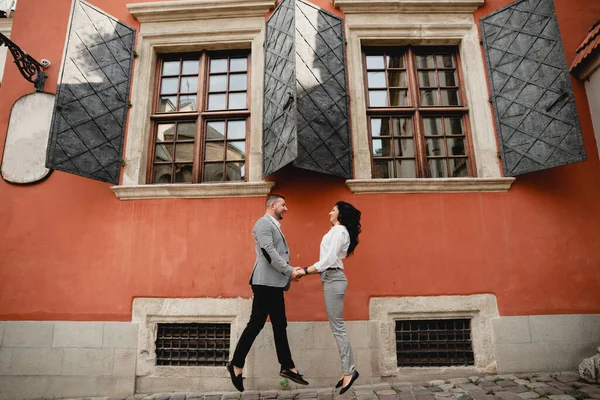 stock image middle-aged couple in love joyfully squeeze together holding hands on the background of a beautiful red old house,young people jump for joy together,couple in love jump together near the house