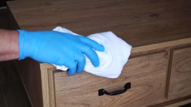 Koruyucu mavi eldivenli bir adam komodinin üzerindeki mobilyaları paçavrayla yıkıyor. Koronavirüsün yayılmasını ve COVID-19 enfeksiyonunu önle. Koronavirüsün yayılmasını nasıl engelleriz?.