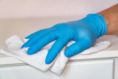 Koruyucu mavi eldivenli bir adam komodinin üzerindeki mobilyaları paçavrayla yıkıyor. Koronavirüsün yayılmasını ve enfeksiyonun COVID-19.