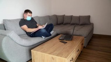 Tıbbi maskeli bir krantin üzerinde bir iş adamı kanepeye oturur ve uzaktan çalışır. Coronavirus COVID19 salgınıyla ilgili evden uzak bir çalışma. Dünya salgını.