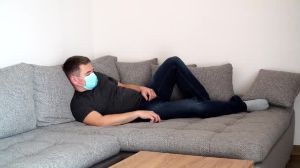 Karantänbunden coronavirus man i medicinsk mask är trött och sover på soffan. Självisolerande pandemi COVID19 — Stockvideo