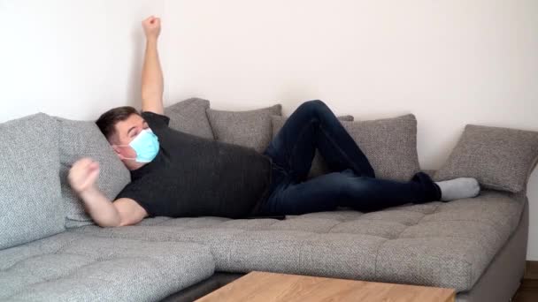 Der unter Quarantäne stehende Coronavirus-Mann in medizinischer Maske breitet sich nach dem Schlafen auf der Couch aus. Selbstisolierungspandemie COVID19 — Stockvideo