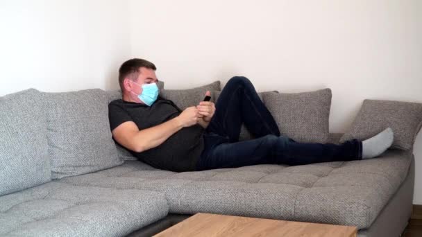 Ein unter Quarantäne stehender Coronavirus-Mann mit medizinischer Maske ist müde und legt sich auf ein Sofa, um am Telefon zu spielen und soziale Netzwerke zu beobachten. Selbstisolierungspandemie COVID19 — Stockvideo