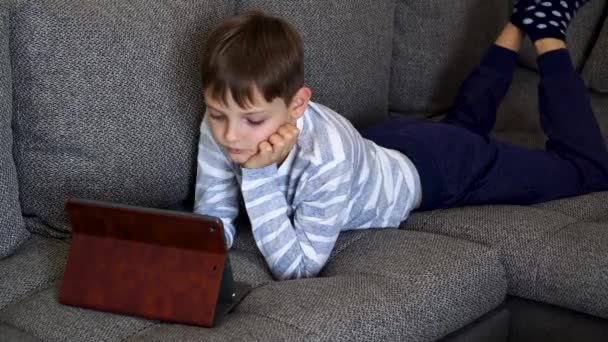 El niño con tableta PC se encuentra en el sofá y los estudios en línea. Lindo caucásico niño educación a distancia en línea — Vídeos de Stock
