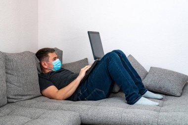 Tıbbi maskeli karantina altındaki bir koronavirüs adamı yoruldu ve dizüstü bilgisayarla çalışmak için kanepeye uzandı. Ev salgını COVID19 'da kendini tecrit karantinası.
