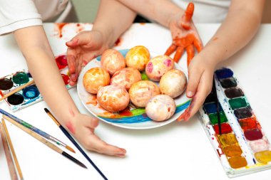 Masanın üzerinde parlak suluboya resimler var. Çocuk elleri yumurta boyar. Paskalya