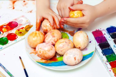 Masanın üzerinde parlak suluboya resimler var. Çocuk elleri yumurta boyar. Paskalya