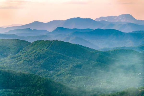 Camadas de montanhas na névoa durante o pôr do sol. Belo pôr-do-sol nas montanhas. Belo pôr do sol em um vale montanhoso com aldeias e neblina nas planícies . — Fotografia de Stock