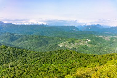 Ufukta evler ve yüksek karlı dağlar ile yeşil bir tepelik vadide gün batımında görünümü.