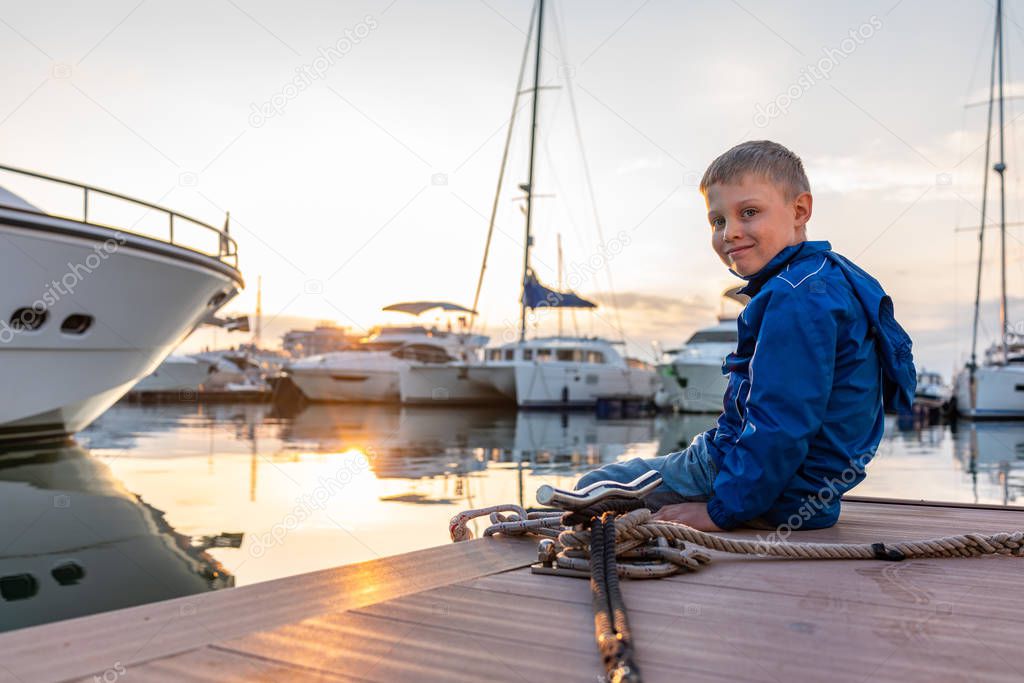 A boy with a smile in a blue jacket sits on a pier with yachts during sunset.