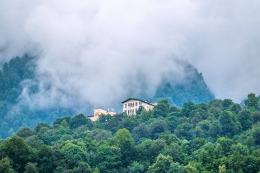 Yeşil ormanların arasındaki dağlardaki otellerin kompleksi. Bulutların ve sislerin arasına gizlenmiş yeşil yamaçlı yüksek dağ. Krasnaya Polyana, Sochi, Rusya