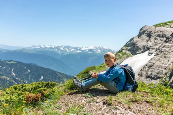 A boy traveler with hiking poles and a backpack is resting in the summer on top of a mountain with snow. Mountain peak in the summer. Caucasus, Russia