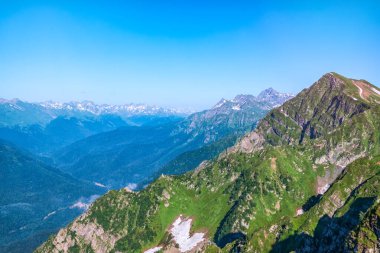 Açık mavi gökyüzüne karşı kayalar ve ağaçlarla kaplı bir dağ sırası. Yaz mevsiminde dağlarda erimemiş kar. Yazın dağ zirvesi. Ufuktaki karla kaplı dağ zirveleri