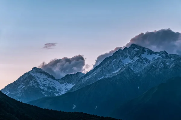 High mountains with snowy peaks at sunset in clouds and fog. High mountains with green slopes and snowy peaks. Mountain ranges in summer sunset