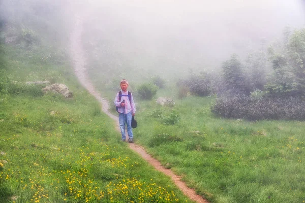 A boy traveler with hiking poles walks along the path on the green slope in heavy fog. The footpath in green field with yellow flowers in dense fog.