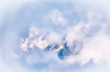 Bulutların ve sislerin arasında karla kaplı yüksek dağ zirvesi. Dağların tepesi bulutlarla çevrili