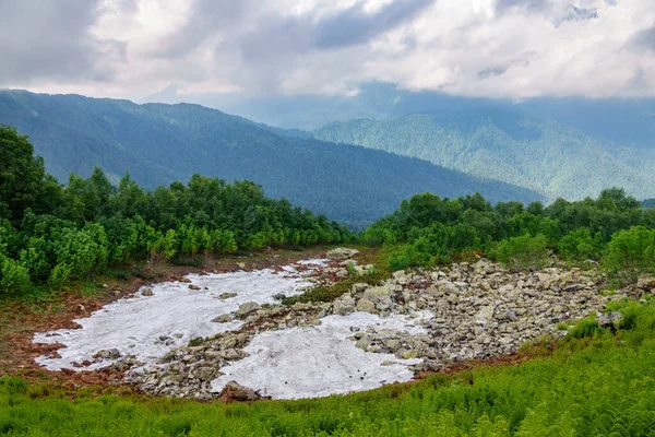 A layer of melting snow in the summer in high mountains. Slopes of mountains are hidden in clouds and fog. Green vegetation in summer.