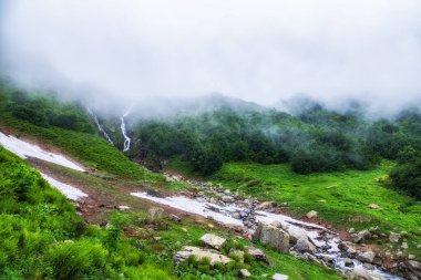 Şelaleli yeşil dağ sıraları ve bahar veya yaz aylarında erimemiş kar. Sis ve bulutlarla kaplı şelaleli güçlü bir dağ nehri akıntısı. Achipsa Şelalesi, Krasnaya Polyana, Sochi, Rusya