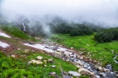 Şelaleli yeşil dağ sıraları ve bahar veya yaz aylarında erimemiş kar. Sis ve bulutlarla kaplı şelaleli güçlü bir dağ nehri akıntısı. Achipsa Şelalesi, Krasnaya Polyana, Sochi, Rusya