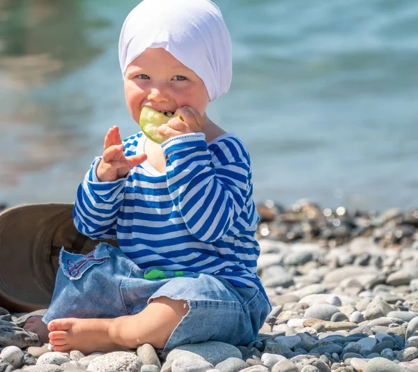 Happy one-year-old child sits on pebbles on the sea beach and eats an apple. Cute baby sits on the coast and eats fruit. Little kid eats outside. Portrait of beautiful baby on blurred sea background.
