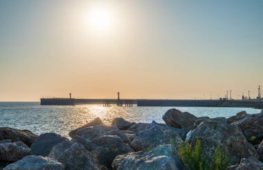 Limanın arka planında büyük taşlar ve kayalar var. Deniz kıyısındaki büyük taşların dalgalanması. Dalgakıran ve deniz feneriyle açık gökyüzü ve parlak gün batımı