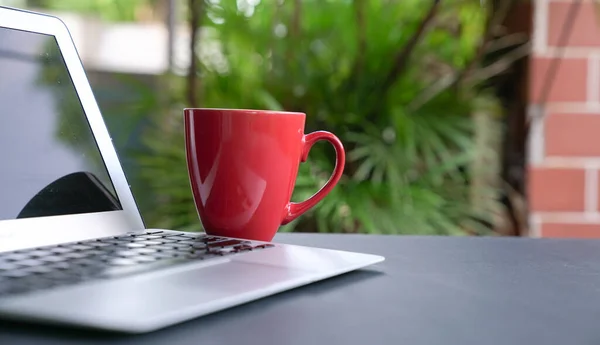 Computer laptop with offee in red cup with green nature background. Work remotely or from home. Selected focus. Copy space.