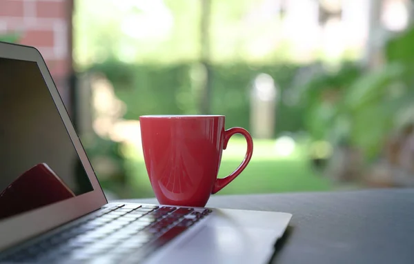 Computer laptop and coffee in red cup with garden background. Work from home. Copy space.