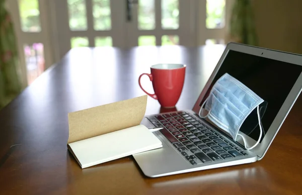 Coronavirus salgını sırasında evde çalış. Bilgisayardaki yüz maskesine ve yemek masasındaki kırmızı kahve fincanına odaklan. Arkaplanda bahçe manzaralı pencereler.