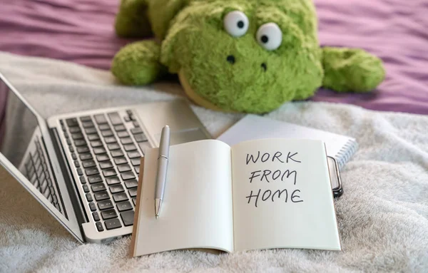 Work from home, text written on notepad. Computer laptop, soft toy and notepad on top of bed in the bedroom.