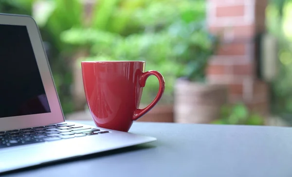 Computer laptop and coffee in red cup with garden background. Work from home. Copy space.