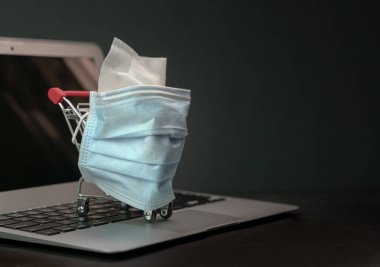 Bilgisayarın üstünde tıbbi maske maskesine sarılı alışveriş arabası ya da alışveriş arabası. Salgın sırasında veya karantina sırasında online alışveriş.