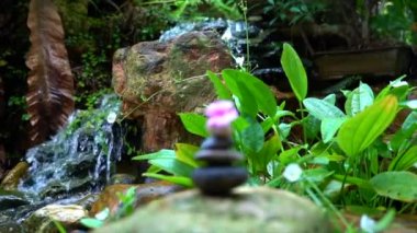 Üstünde pembe bir çiçek olan çakıl taşları piramidin içine istiflenmiş. Arka planda küçük şelale ve kaya bahçesi var. Zen, spa, güzellik ve sağlık konsepti