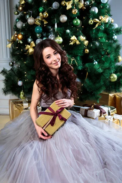 Bela jovem mulher em vestido de noite de luxo detém uma caixa de presente em um fundo de árvore de natal — Fotografia de Stock