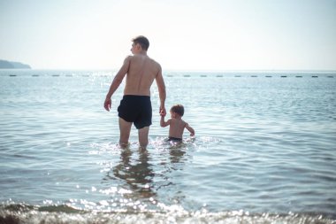 Genç baba ve küçük oğlu suda birlikte oynuyorlar. Manzaralı deniz ve dağ manzarası, yaz tatili