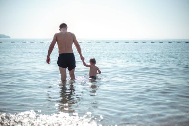 Genç baba ve küçük oğlu suda birlikte oynuyorlar. Manzaralı deniz ve dağ manzarası, yaz tatili