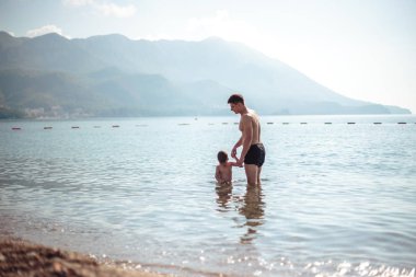 Genç baba ve küçük oğlu suda birlikte oynuyorlar. Manzaralı deniz ve dağ manzarası, yaz tatili