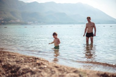 Genç baba ve küçük oğlu suda birlikte oynuyorlar. Manzaralı deniz ve dağ manzarası, yaz tatili