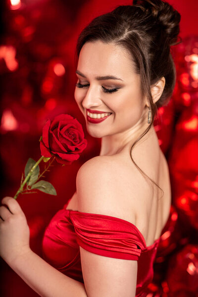 Beautiful young woman in red evening dress posing over red background with big heart shape balloons. 