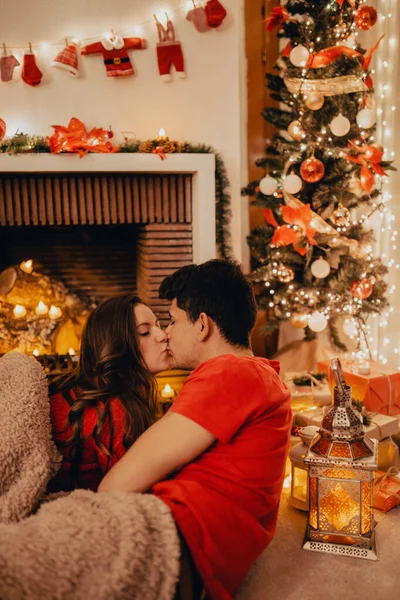 คู่รักที่หลงใหลฉลองคริสต์มาสด้วยกัน หญิงสาวและชายจูบและกอดกันใต้เตาผิงพร้อมเทียนของขวัญและต้นไม้ที่ตกแต่ง สุขสันต์วันคริสต์มาสและปีใหม่ . — ภาพถ่ายสต็อก