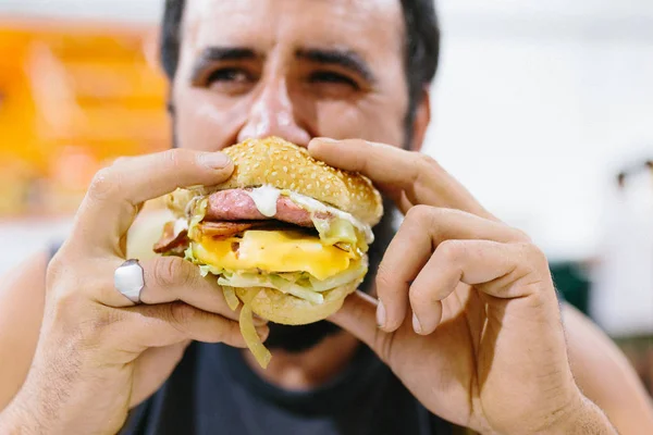 Zblízka portrét vousatého muže, jak jí burger. Portrét expresivního tlusťocha, jak jí hamburger s masem, rajčaty, slaninou, cibulí a sýrem. Cheeseburger a fast food koncept. — Stock fotografie