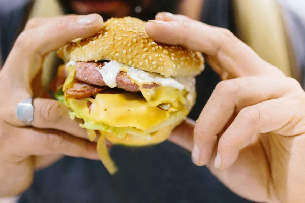 Zblízka portrét vousatého muže, jak jí burger. Portrét expresivního tlusťocha, jak jí hamburger s masem, rajčaty, slaninou, cibulí a sýrem. Cheeseburger a fast food koncept. — Stock fotografie