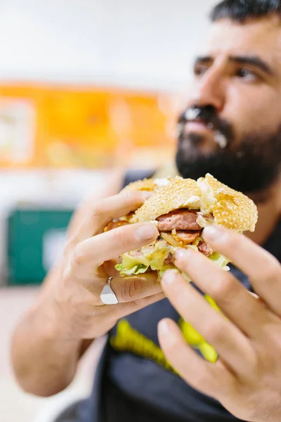 Zblízka portrét vousatého muže, jak jí burger. Portrét expresivního tlusťocha, jak jí hamburger s masem, rajčaty, slaninou, cibulí a sýrem. Cheeseburger a fast food koncept. — Stock fotografie